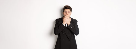Scared man in formal suit, gasping and looking frightened at camera, standing against white...