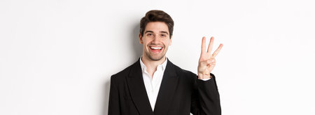 Close-up of handsome businessman in black suit, smiling amazed, showing number three, standing over...