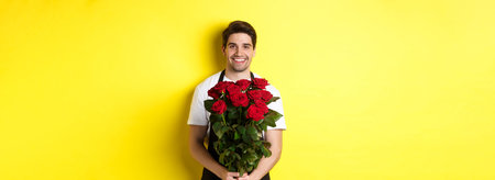 Smiling florist in black apron holding flowers, selling bouquet of roses, standing over yellow...