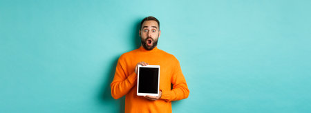Technology. Surprised bearded man drop jaw, staring at camera and showing digital tablet screen, standing again light blue backgroundの写真素材