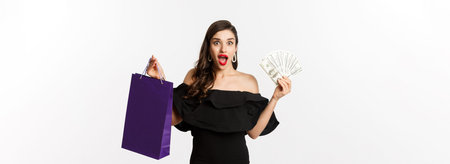 Happy woman buyer holding shopping bag and money, standing in black dress over white background
