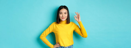 Beautiful smiling asian woman recommend product, showing Ok sign and looking satisfied, standing...