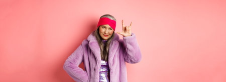 Funny and cool asian senior woman showing rock n roll gesture, looking sassy, standing over pink...