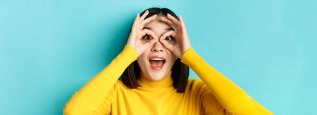 Close up of funny asian woman looking through hand binoculars with surprised face, see something...
