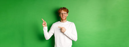 Handsome young man with ginger hair and glasses, pointing fingers right at copy space and smiling, standing over green backgroundの写真素材