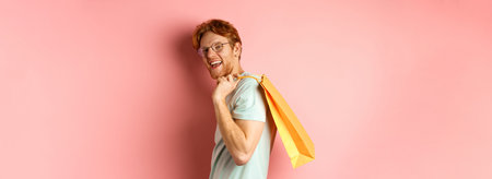 Carefree young man with red hair and glasses, walking with shopping bag over his shoulder and...