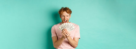 Excited young man winning prize money, counting cash and looking amazed at dollars, standing over...