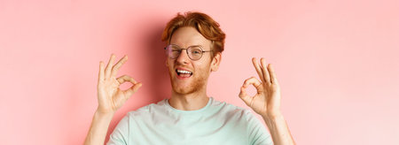Cheeky redhaed guy in glasses assuring you, winking and showing okay signs, guarantee good quality,...