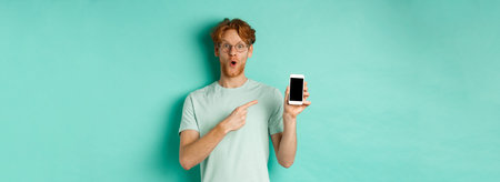Check this out. Handsome redhead guy in glasses pointing finger at blank smartphone screen, showing...