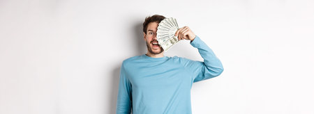 Surprised and happy young man covering half of face with money, gasping amazed at camera, checking...