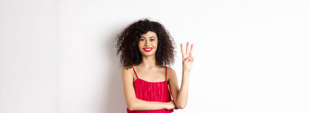 Beautiful lady with curly hair, wearing elegant red dress and showing number three, making order,...