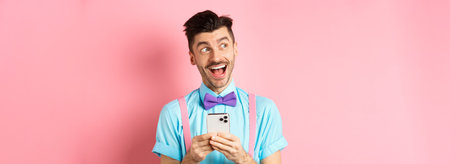 Technology concept. Excited man looking aside with dreamy face, winning online prize on smartphone, standing cheerful over pink backgroundの写真素材