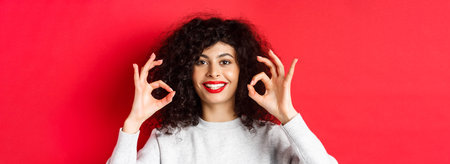 Close-up portrait of smiling woman with curly hair and red lips, showing okay gesture and looking...