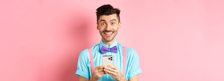 Online shopping. Happy guy in bow-tie, smiling at camera and using smartphone, standing on pink...