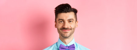 Close-up of smiling hansome man with moustache, wearing bow-tie and shirt, standing cheerful on pink...