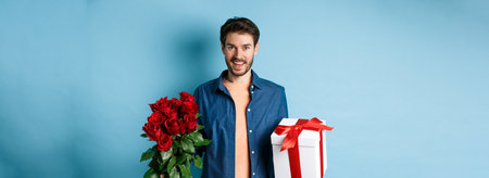 Love and relationship concept. Happy young man bring flowers and gift on romantic date. Boyfriend wish bouquet of roses and present standing on blue backgroundの写真素材