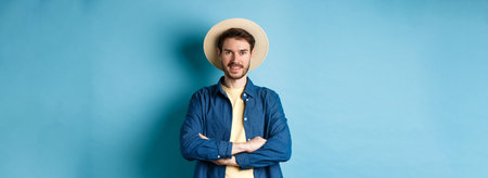 Portrait of cheerful guy going on vacation in straw summer hat, cross arms on chest and smiling, looking confident, standing on blue backgroundの写真素材