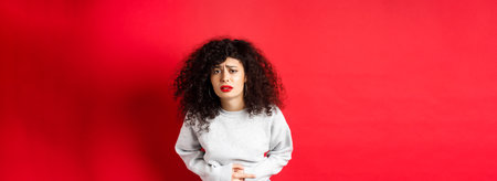 Image of young woman having stomach ache, bending from pain and complaining on painful menstrual cramps, standing on red backgroundの写真素材