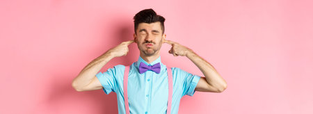 Annoyed funny guy in bow-tie shut ears, plug fingers and grimacing, irritated by loud noise,...