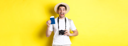 Travelling, vacation and tourism concept. Excoted tpirost showing passport with tickets, holding camera and wearing straw hat, standing over yellow backgroundの写真素材