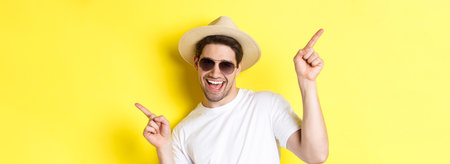 Concept of tourism and vacation. Close-up of man enjoying holidays on trip, dancing and pointing fingers sideways, wearing sunglasses with straw hatの写真素材