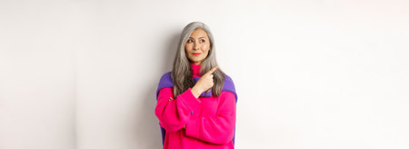 Stylish modern asian elderly woman pointing at upper left corner, looking at logo with pleased smile, standing in pink sweater over white backgroundの写真素材
