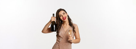 Winter holidays celebration concept. Happy woman on Christmas party saying cheers, raising bottle of champagne and smiling, standing in elegant dress, white backgroundの写真素材