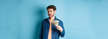 Unbothered rich guy showing cash and smiling, holding dollars in hand, standing on blue background