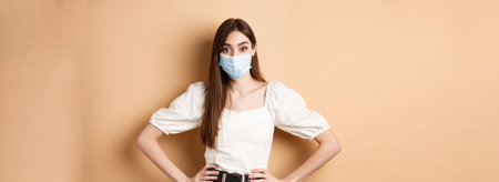 Woman in medical mask looking alarmed at camera, standing on beige background