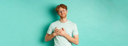 Handsome young man with red hair and glasses, holding hands on heart and smiling, saying thank you, feeling grateful and touched, standing over turquoise backgroundの写真素材
