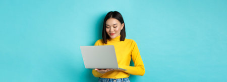 Beautiful asian female student working on laptop, typing on keyboard and looking at screen with...