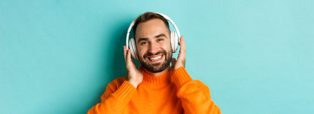 Close-up of handsome modern man listening music in headphones, standing in orange sweater over...