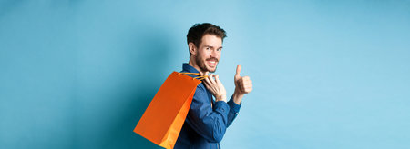 Cheerful shopper holding orange shopping bag on shoulder, turn around at camera with thumbs up,...