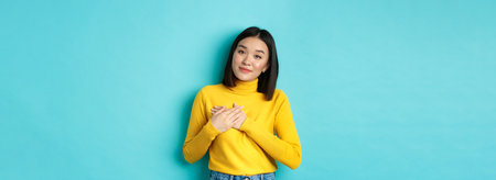 Portrait of beautiful heartfelt woman holding hands on heart, smiling and listening compassionate, standing over blue background in yellow pulloverの写真素材