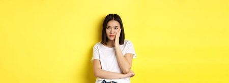 Bored and annoyed asian woman tired of listening, looking skeptical at camera, standing over yellow...