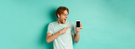 Check this out. Handsome redhead guy in glasses pointing finger at blank smartphone screen, showing...
