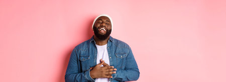 Happy Black hipster guy in beanie and denim shirt, laughing out loud, touching stomach and smiling...