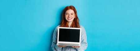 Young smiling woman with red hair and freckles showing computer screen, holding laptop and demonstrate online promo, standing over blue backgroundの写真素材