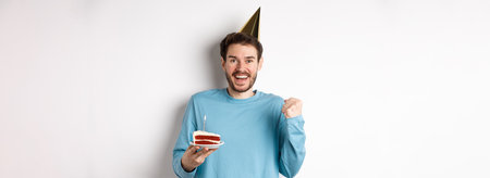 Celebration and holidays concept. Cheerful young man celebrating birthday in party hat, saying yes and fist pump in joy, holding bday cake, white backgroundの写真素材