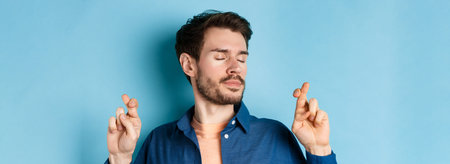 Close up of hopeful young man cross fingers for good luck and close eyes, making wish with calm...