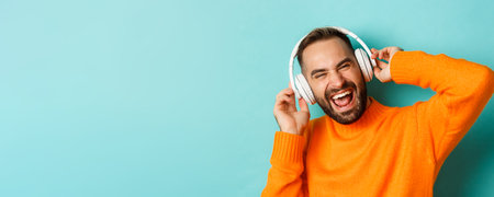 Close-up of handsome modern man listening music in headphones, standing in orange sweater over turquoise backgroundの写真素材