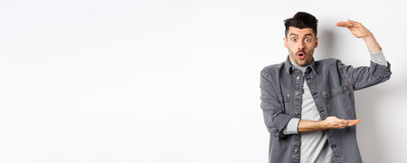 Cheerful guy smiling and showing big size, showing large thing and looking excited, standing against white backgroundの写真素材