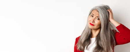 Close up portrait of confused asian female manager scratching head and looking questioned at camera,...