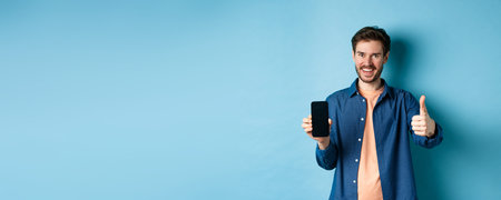 Handsome young man showing thumbs up gesture and empty smartphone screen, recommending app or...