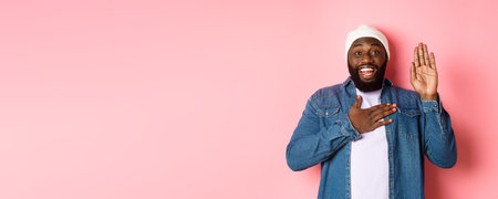 Image of smiling african-american man being honest, telling truth, hands on heart and in air, making promise or swearing, standing ove rpink backgroundの写真素材