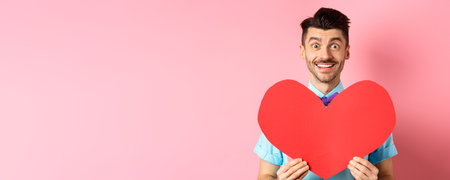 Hopeful man in love showing red heart sign, smiling at camera, waiting for soulmate on Valentines day, standing on pink backgroundの写真素材