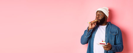 Happy Black hipster celebrating birthday, blowing party whistle, holding bday cake with candle,...