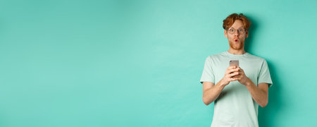 Impressed young man with red hair and glasses, holding mobile phone, saying wow and stare amazed at camera, standing over mint backgroundの写真素材