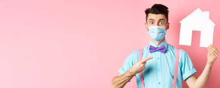 Covid, pandemic and real estate concept. Amazed man in face mask pointing at paper house cutout, standing impressed on pink backgroundの写真素材