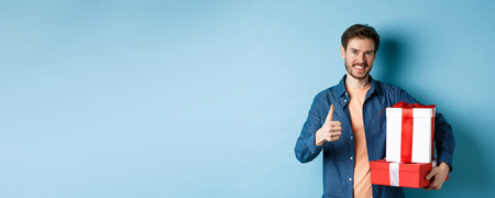 Smiling man holding romantic gifts and showing thumbs-up, celebrating Valentines day, buying presents for lover, standing over blue backgroundの写真素材